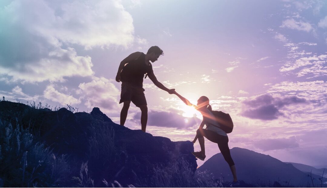 One hiker helping another climb a mountain, symbolising how intentional financial planning can help build and protect long-term wealth.