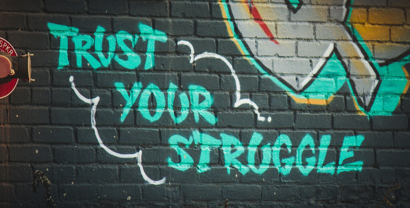 Graffiti on a brick wall displaying the words “Trust Your Struggle”, symbolising personal growth, resilience and the value of overcoming challenges.