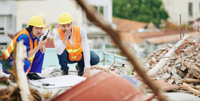 Two people in safety helmets and high-visibility vests inspecting damaged property and reviewing plans, illustrating the importance of protecting assets and preparing for unexpected setbacks.
