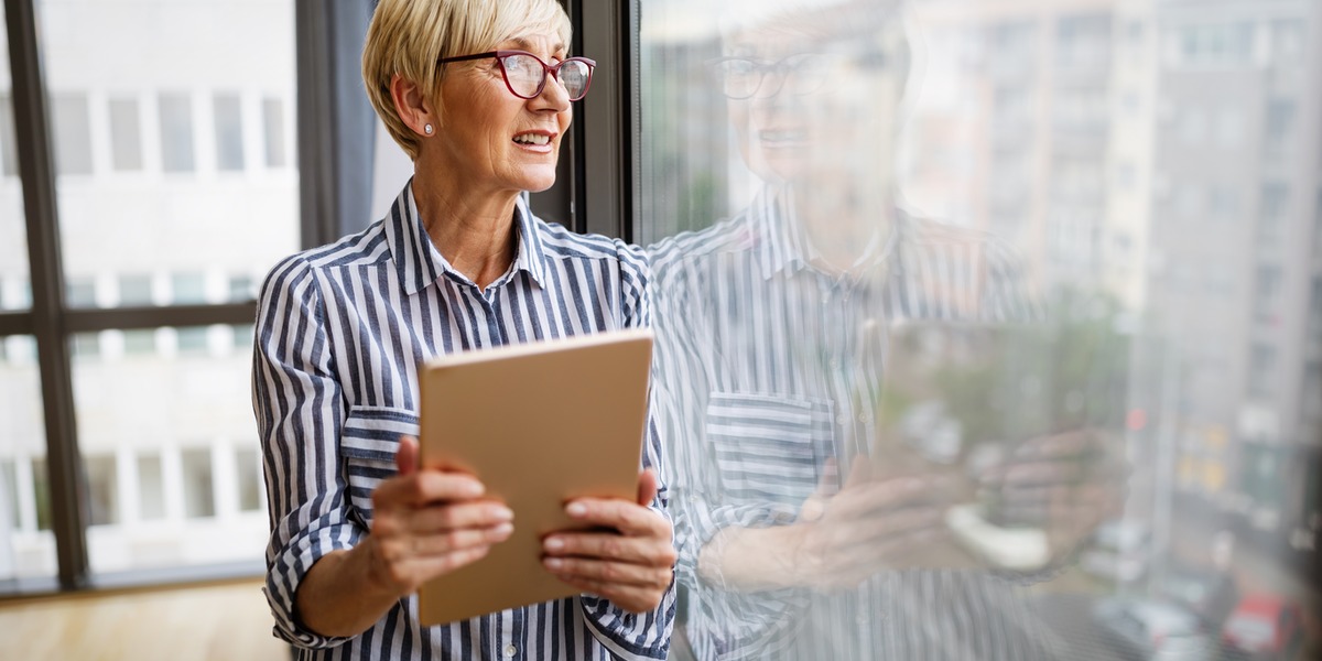 Older professional holding a tablet beside an office window, illustrating meaningful work and new opportunities during the productive retirement years between 50 and 80.